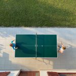 Table tennis table from above, with two players, on a sealed surface, with a building and a lawn.