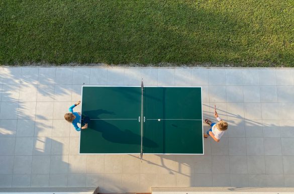Tischtennisplatte von Oben aufgenommen, mit zwei Spielern, auf versiegeltem Untergrund, mit Gebäude und Rasen.