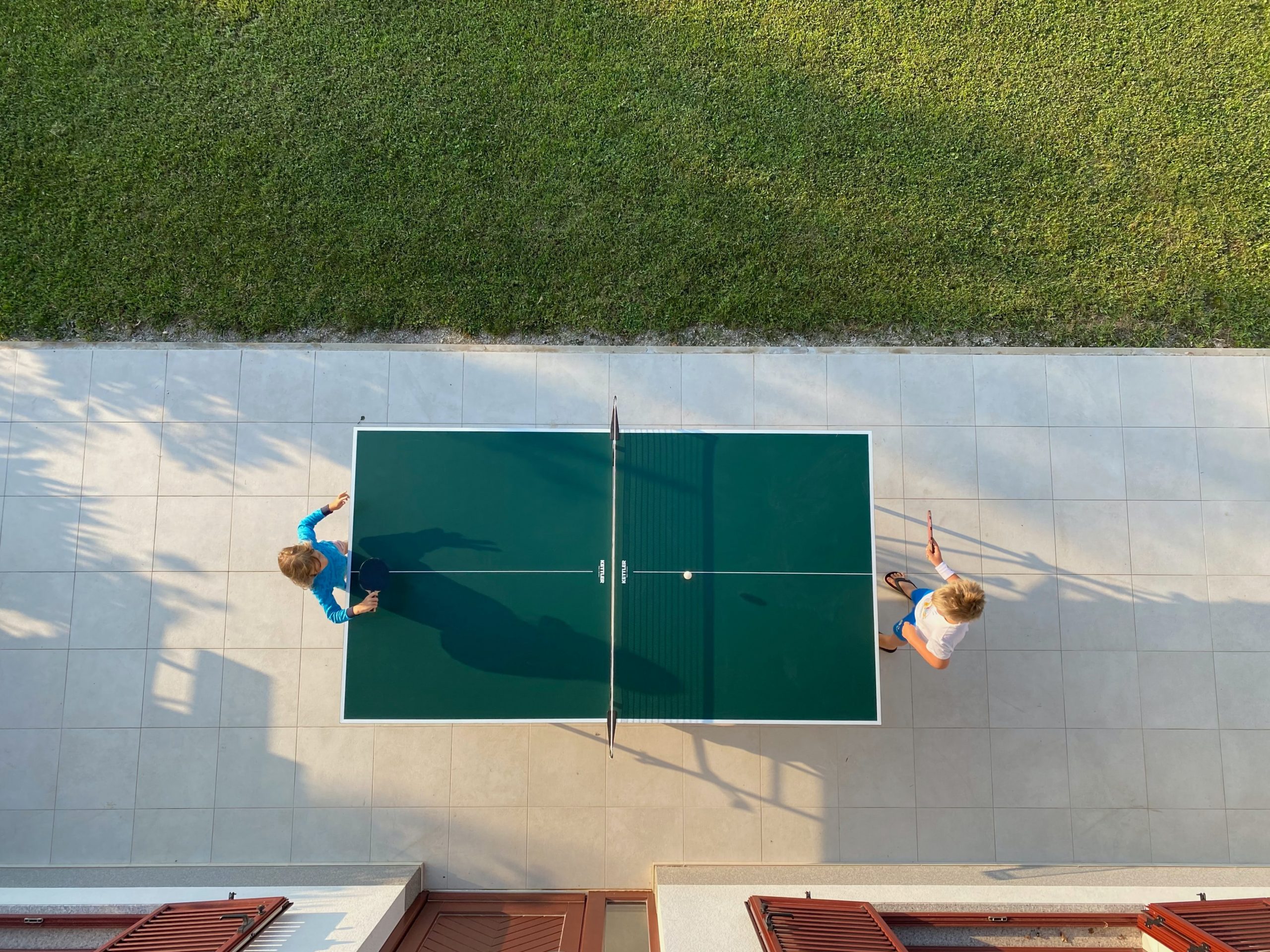Table tennis table from above, with two players, on a sealed surface, with a building and a lawn.